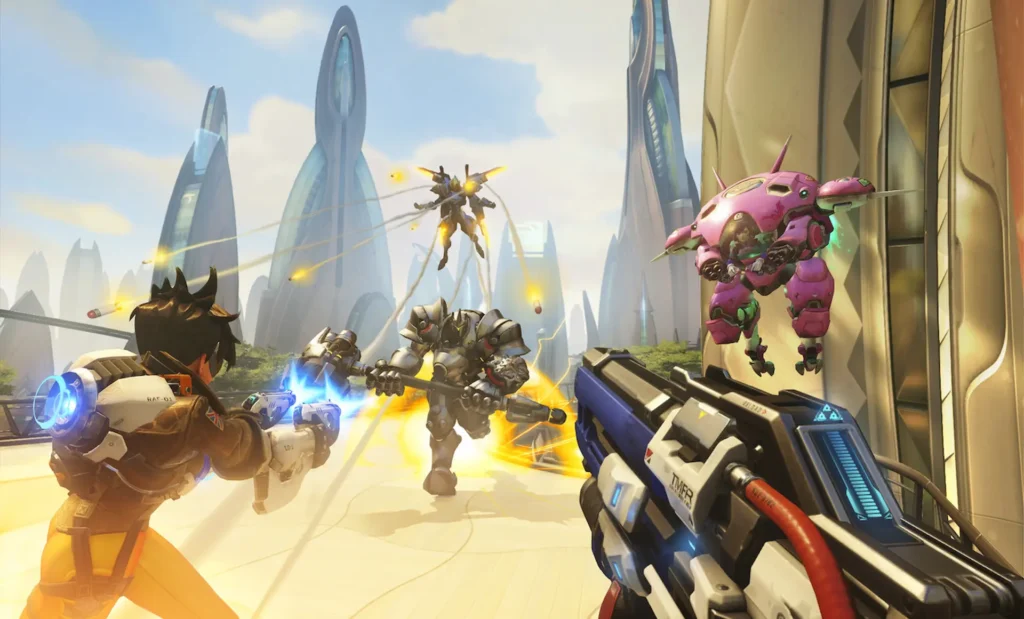 Vista en primera persona de Tracer disparando a Reinhardt, mientras D.Va y Pharah vuelan en una partida de Overwatch.