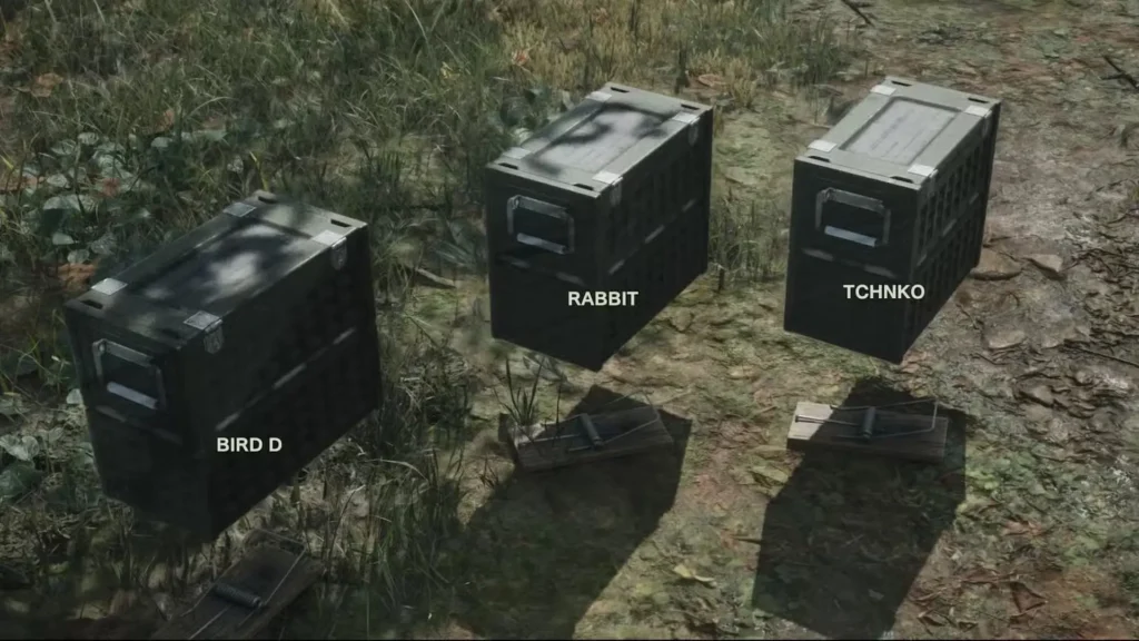 En la imagen, se muestran tres cajas metálicas de aspecto militar, con las etiquetas "BIRD D", "RABBIT" y "TCHNKO" respectivamente, apiladas sobre la hierba junto a dos trampas de madera.