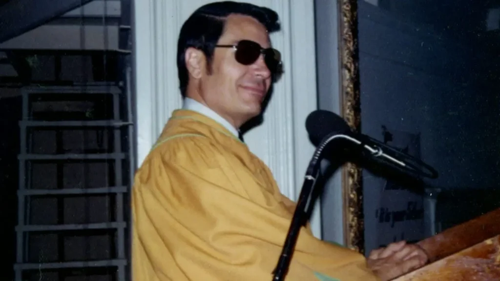 Hombre con gafas de sol y bata amarilla sonríe mientras habla al micrófono en un púlpito.