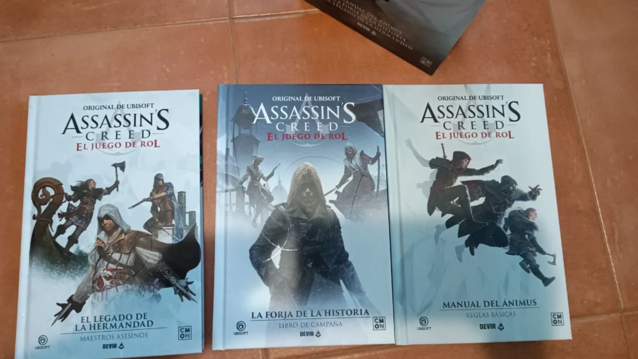 Las portadas de los tres libros del juego de rol de Assassin's Creed se exhiben sobre un suelo de baldosas, destacando a diversos asesinos de la franquicia.