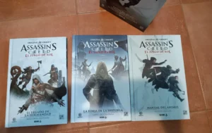 Las portadas de los tres libros del juego de rol de Assassin's Creed se exhiben sobre un suelo de baldosas, destacando a diversos asesinos de la franquicia.