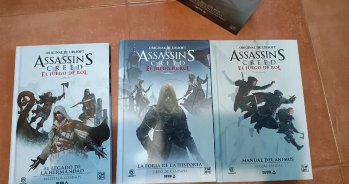 Las portadas de los tres libros del juego de rol de Assassin's Creed se exhiben sobre un suelo de baldosas, destacando a diversos asesinos de la franquicia.