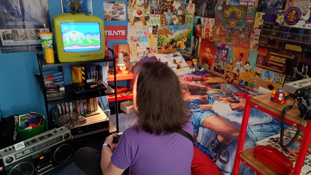 Un chico jugando al Sonic en una sala decorada al estilo 80 con muchísimas referencias a juguetes, series y tecnología de la época.