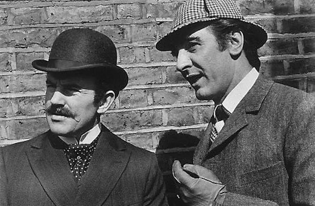 Sherlock Holmes, con gorra de cazador, y el doctor Watson, con bombín, charlan ante una pared de ladrillo